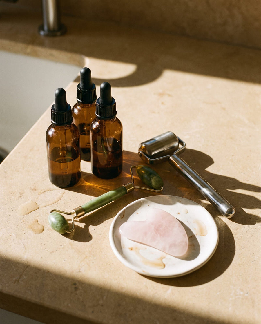 Three dropper bottles, a jade roller, a steel roller and a gua sha stone on a bathroom counter